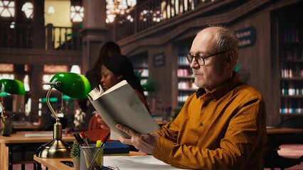 Elderly man immersed in reading and writing at a university campus library, working on his thesis with intellectual ambition and dedication to learning and academic achievement.