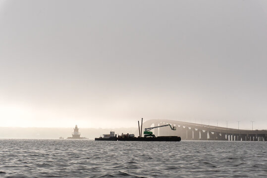 Construction Barge and Crane on Foggy Bay with Historic Lighthouse and Long Bridge in Background