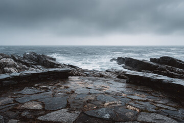 Obraz premium A rugged stone pier extends into the turbulent sea under a grey, misty sky, waves crashing against the rocks in a cold, dramatic atmosphere.