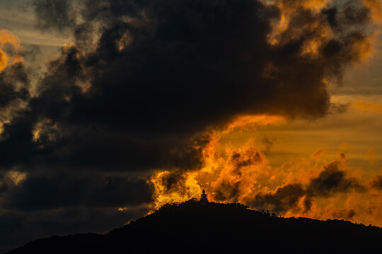 The golden sun sets behind the mountain casting a warm glow around the grand Big Buddha of Phuket. Silhouetted against a radiant sky, the sacred statue radiates peace and spiritual serenity.