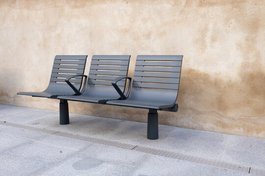 Modern gray bench with armrests positioned against a textured beige wall, showcasing minimalist design and urban seating for public spaces and outdoor environments