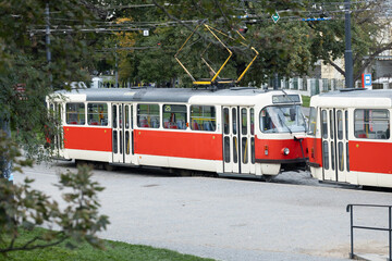 Naklejka premium Vintage red and white tram is navigating through a city street surrounded by greenery, showcasing urban transportation and historical architecture in a vibrant setting