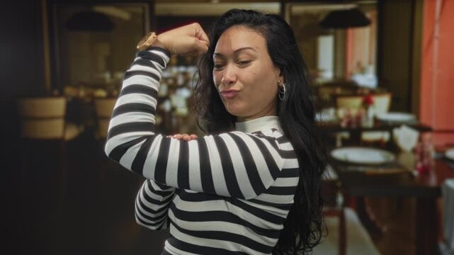 Woman flexes arm and poses with fist at forehead in restaurant dining room; confidence empowerment pride.