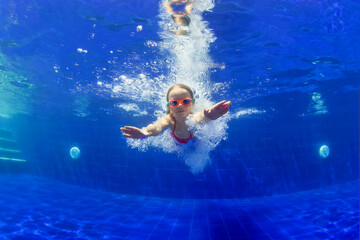 Underwater shot of happy smiling young child jumping with splashes and diving with fun in blue swimming pool. Healthy family lifestyle, water sport activity, swimming lessons on holidays with kids