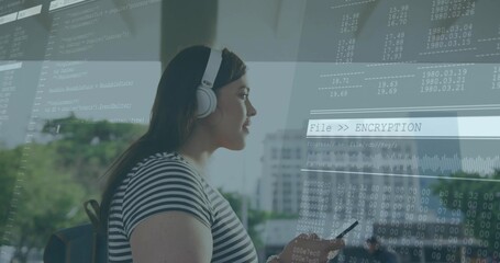 Standing woman wearing headphones holding smartphone near glass panel with code overlay, copy space