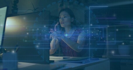 Woman in burgundy top calling smartphone and interacting with holographic interface in night office