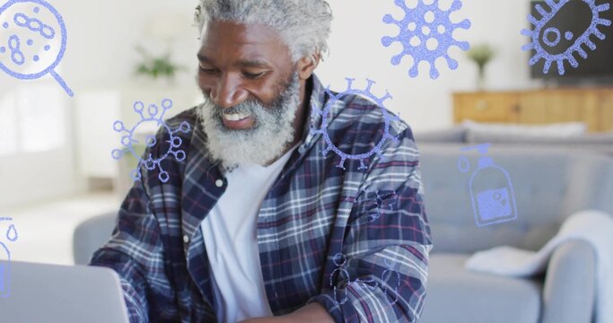 Smiling senior African American man focusing on laptop at home, with virus icons and sanitizer - Powered by Adobe