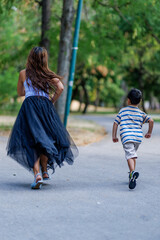 Rear view of a woman and a child running down a paved pathway surrounded by lush green park scenery