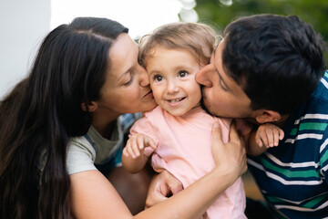 Loving parents warmly embrace and kiss their cheerful toddler outside in a cozy family moment.