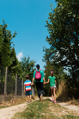 Obraz premium Mother walking outdoors on a dirt trail holding hands with her two children, enjoying a sunny day surrounded by lush nature and clear skies.