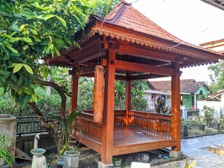 A beautifully carved traditional Javanese wooden gazebo, called a "pendopo" or "limasan", stands outdoors surrounded by lush green trees and plants.
