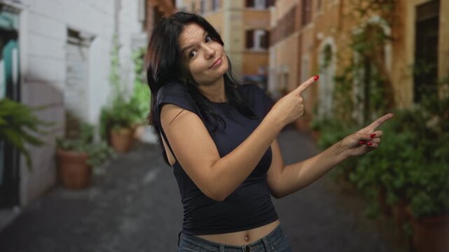 Woman with exposed navel points finger to side on sunlit street between pastel buildings; curiosity.
