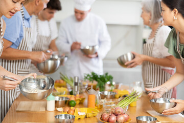 Cooking course - senior male chef in cook uniform teaches young people cooking class students to prepare and mix ingredients for dishes in restaurant kitchen