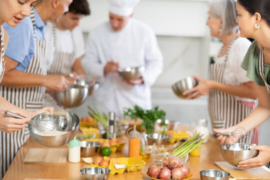 Cooking course - senior male chef in cook uniform teaches young people cooking class students to prepare and mix ingredients for dishes in restaurant kitchen