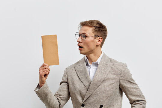 Professional man in beige suit and glasses holding blank card, looking surprised or puzzled, studio portrait on plain gray background, concept of communication, business, presentation.