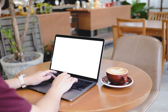 A person's hands typing on a laptop with a blank white screen in an outdoor cafe setting, with a cup of coffee on the table.