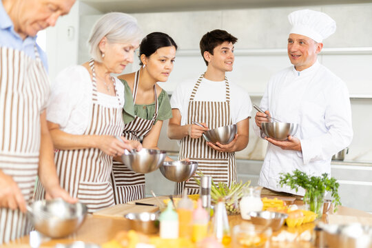 Friendly experienced male chef in white uniform giving culinary classes, teaching to cook to group of men and women of different ages.. - Powered by Adobe