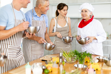 Smiling old woman chef of cooking course teaching attendees how to mix sauce using whisk and bowl