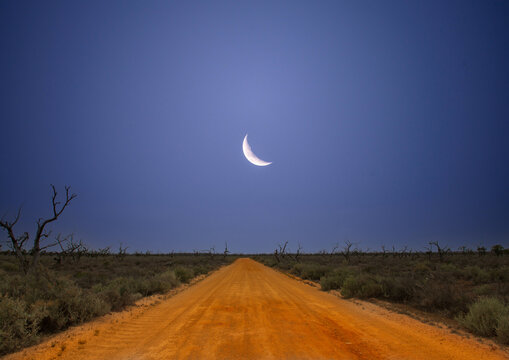 Desert road in outback Australia with moon in blue sky