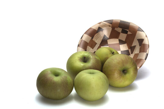 Green apples spilling out of a wooden bowl. The apples are in the foreground and the bowl is in the back.  The composition is on a white background.  A very clean and simple vibe.