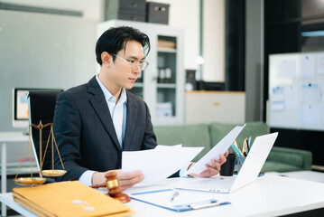 Professional Asian lawyer reading legal documents in modern office with gavel and scales on desk.