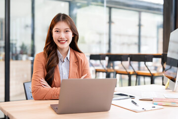 Professional young Asian businesswoman smiling confidently while working on a laptop in a modern office or coworking space.