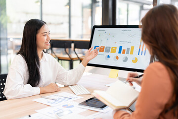 Two Asian colleagues or businesswomen analyzing financial charts and data on a desktop monitor, collaborating on a project in a modern office.