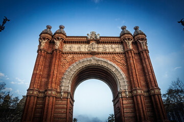 Obraz premium Low-angle view of the Arc de Triomf in Barcelona, Spain. The red brick monument with sculpted friezes and large arch stands against a clear blue sky, highlighting the Neo Mudejar civic gateway.