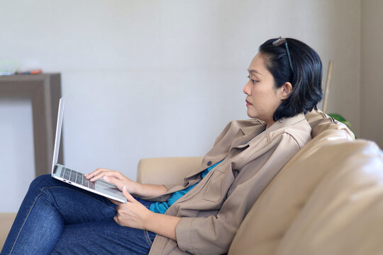Workspace, side view of Asian woman sitting on sofa, using laptop for online business, chatting, negotiating or working from home - Powered by Adobe
