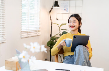 Smiling and relaxed young Asian woman enjoying a coffee break while pausing from work on her digital tablet, bathed in natural light from a window in a modern home office.