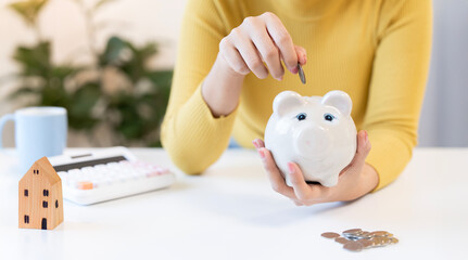 Close-up of a young Asian woman's hands putting a coin into a white piggy bank, symbolizing savings, personal finance, and long-term investment planning, with a calculator and house model on the desk.