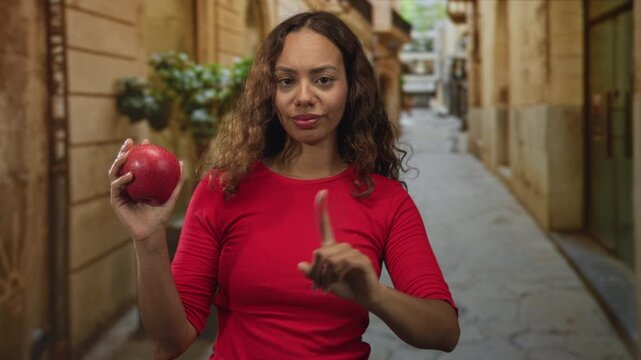 Young woman holding red apple and points finger in a narrow city street scene; confidence healthy eating.