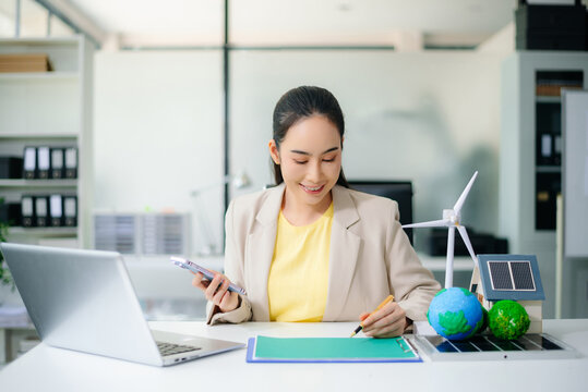 Asian businesswoman in office planning green energy with wind turbine, solar panel, globe. Concept of sustainability, climate change, and reducing CO2 emissions. - Powered by Adobe