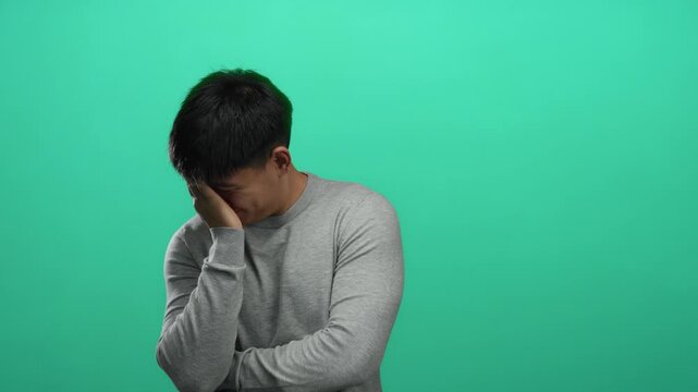 Young man in grey sweater with hand on face over isolated green background exuding contemplation and calmness, blending softly into the vibrant backdrop.