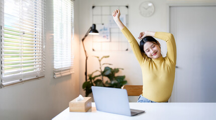 A cheerful young Asian woman in a yellow sweater stretches her arms above her head with a satisfied smile while taking a break from working on her laptop at a white desk in a brightly lit home office,