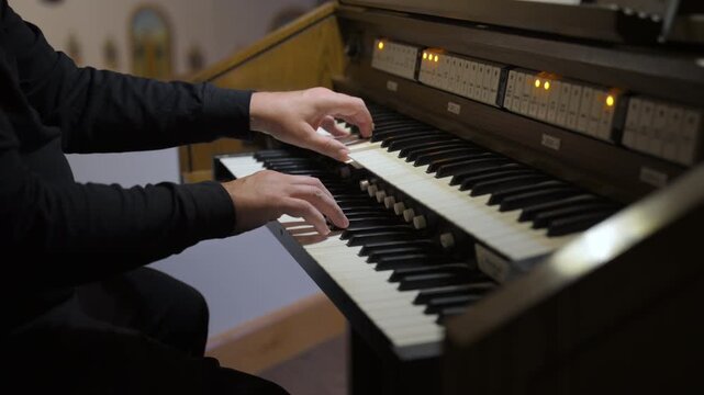 Musician playing an organ or inside a church or concert hall. The performer hands are actively moving across multiple manuals keyboards, suggesting live performance. 