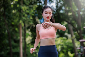 Fototapeta premium Asian woman running in a park, checking her smartwatch for workout progress. Maintaining healthy lifestyle and fitness
