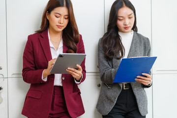 Two Asian businesswomen analyzing data, using a digital tablet and clipboard, standing in a modern office environment