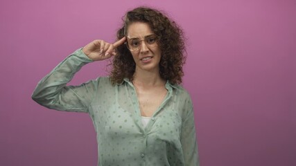 Woman points finger and grimaces in studio against pink wall, wearing green polka dot blouse with glasses and curly hair; frustration.