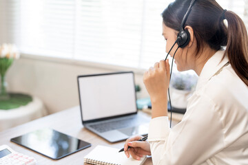 A focused young Asian female call center agent wearing a headset, speaking on the microphone, and taking notes while providing customer support at her desk.