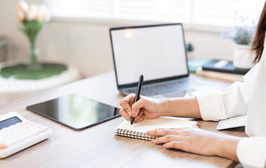 Close-up of a businesswoman's hands writing notes in a spiral notebook with a pen at her office desk, next to a laptop and calculator.