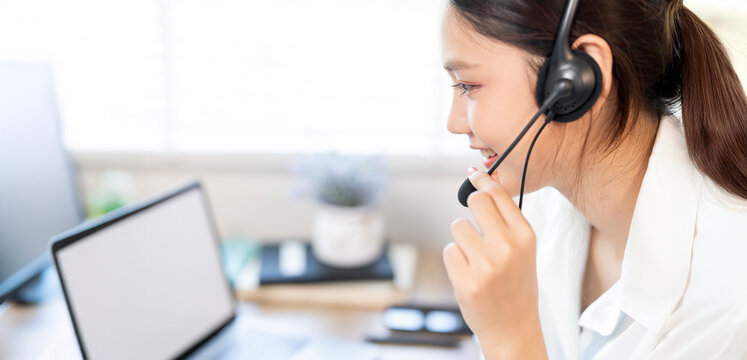 A young, smiling Asian female call center agent or customer service representative wearing a headset and talking with a customer at her desk.