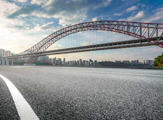 Obraz premium Empty asphalt road with a large red arch bridge and modern city skyline in the background.