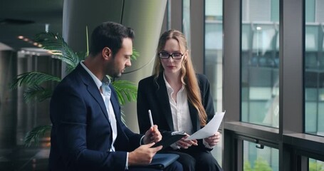 Professional businessman and businesswoman collaborate reviewing documents and using digital tablet during meeting in modern office lobby near window - Powered by Adobe
