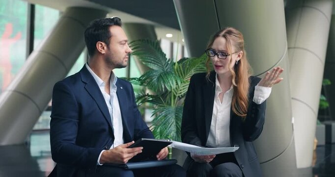 Bossy businesswoman looks unhappy gesturing while giving critical feedback on documents to male employee holding tablet during meeting in modern office lobby