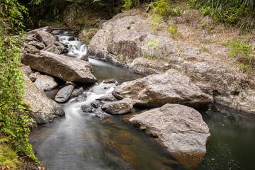 Large granite boulders litter the floor of a stream in this long exposure landscape of cascades and pools surrounded by rainforest at Crystal Cascades in Cairns in tropical North Queensland, Australia
