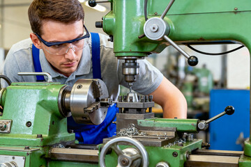 Focused young male machinist or metalworker wearing safety glasses operating an industrial green lathe machine, drilling or turning a metal workpiece covered in shavings