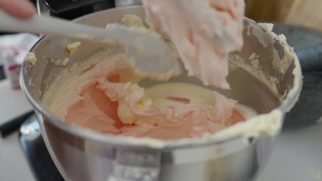 A baker lifts the paddle and scraps the inside of a mixer bowl with whipped buttercream with a spatula and gently taps it on the side and continues to scraps the sides.