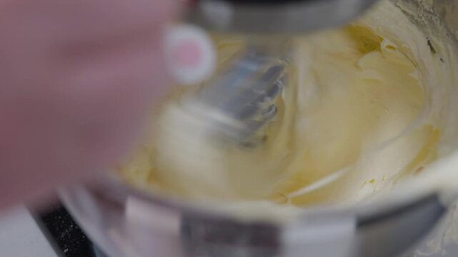 Close up, A baker adds drops of pink food colouring into whipping buttercream frosting as the paddle spins around.