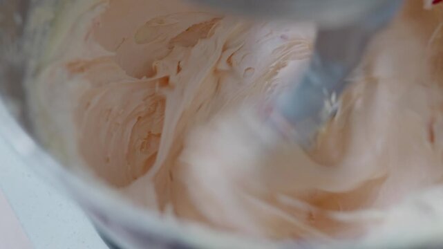 Close up, A cook drops food colouring into pink buttercream icing whipping in a stand mixer bowl on high speed.